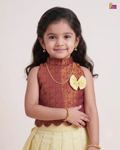 A young girl modeling a traditional Indian outfit featuring a sleeveless maroon top with gold floral embroidery, a yellow bow accent, and a yellow skirt, set against a plain light background.