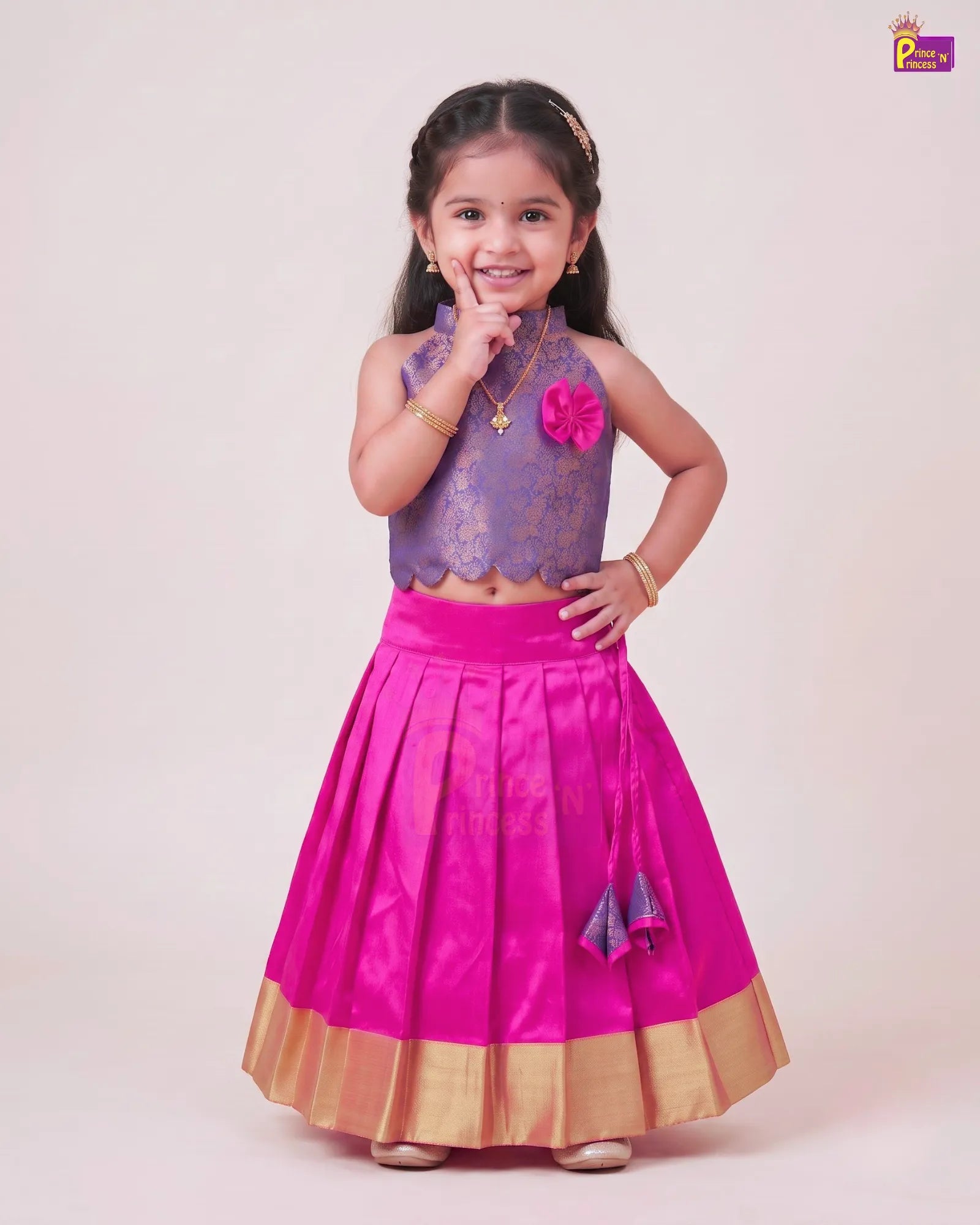 A young girl modeling a traditional Indian outfit consisting of a purple lace sleeveless crop top with a pink flower accent and a bright pink pleated skirt with a gold border, set against a plain beige studio background.