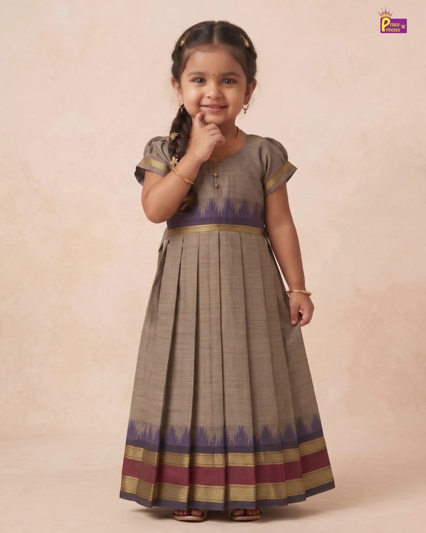 A young girl modeling a traditional Indian salwar kameez dress. The dress features a taupe-beige body with a purple and gold ikat border at the waist and hem, paired with a matching pleated skirt.