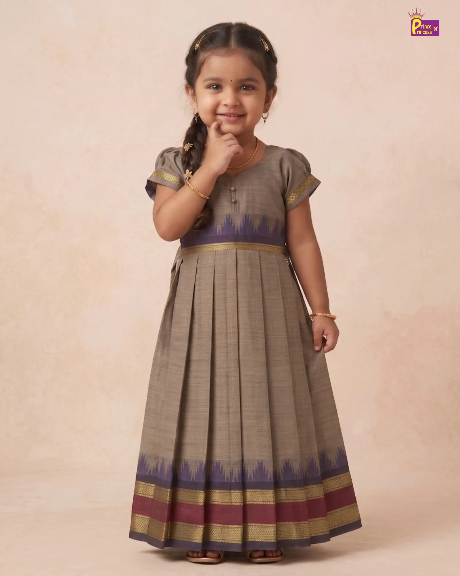 A young girl modeling a traditional Indian salwar kameez dress. The dress features a taupe-beige body with a purple and gold ikat border at the waist and hem, paired with a matching pleated skirt.