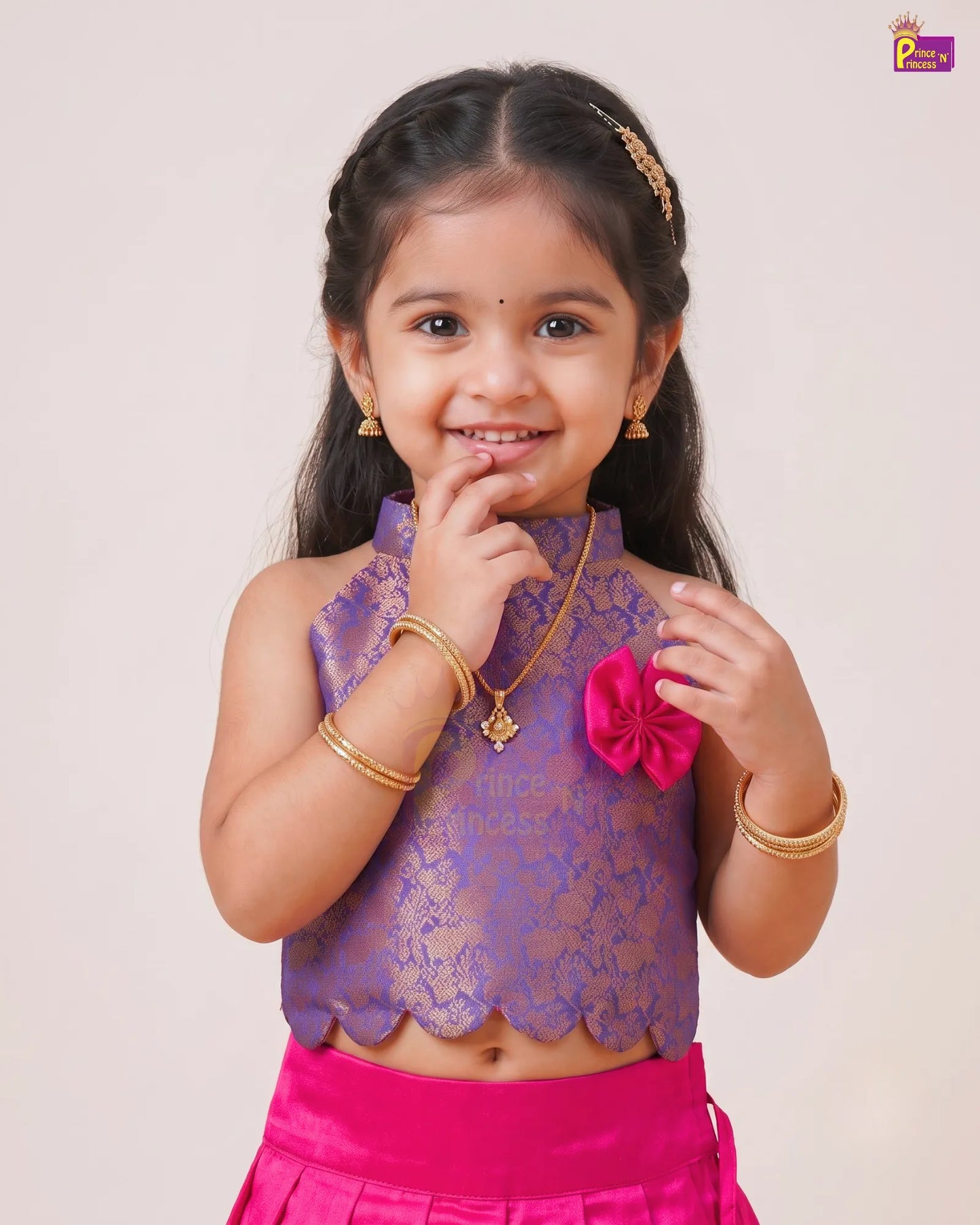 A young girl modeling a traditional Indian outfit featuring a purple lace sleeveless top with gold embroidery, a bright pink skirt, and gold jewelry, set against a plain light beige background.
