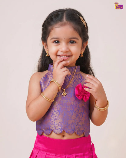 A young girl modeling a traditional Indian outfit featuring a purple lace sleeveless top with gold embroidery, a bright pink skirt, and gold jewelry, set against a plain light beige background.