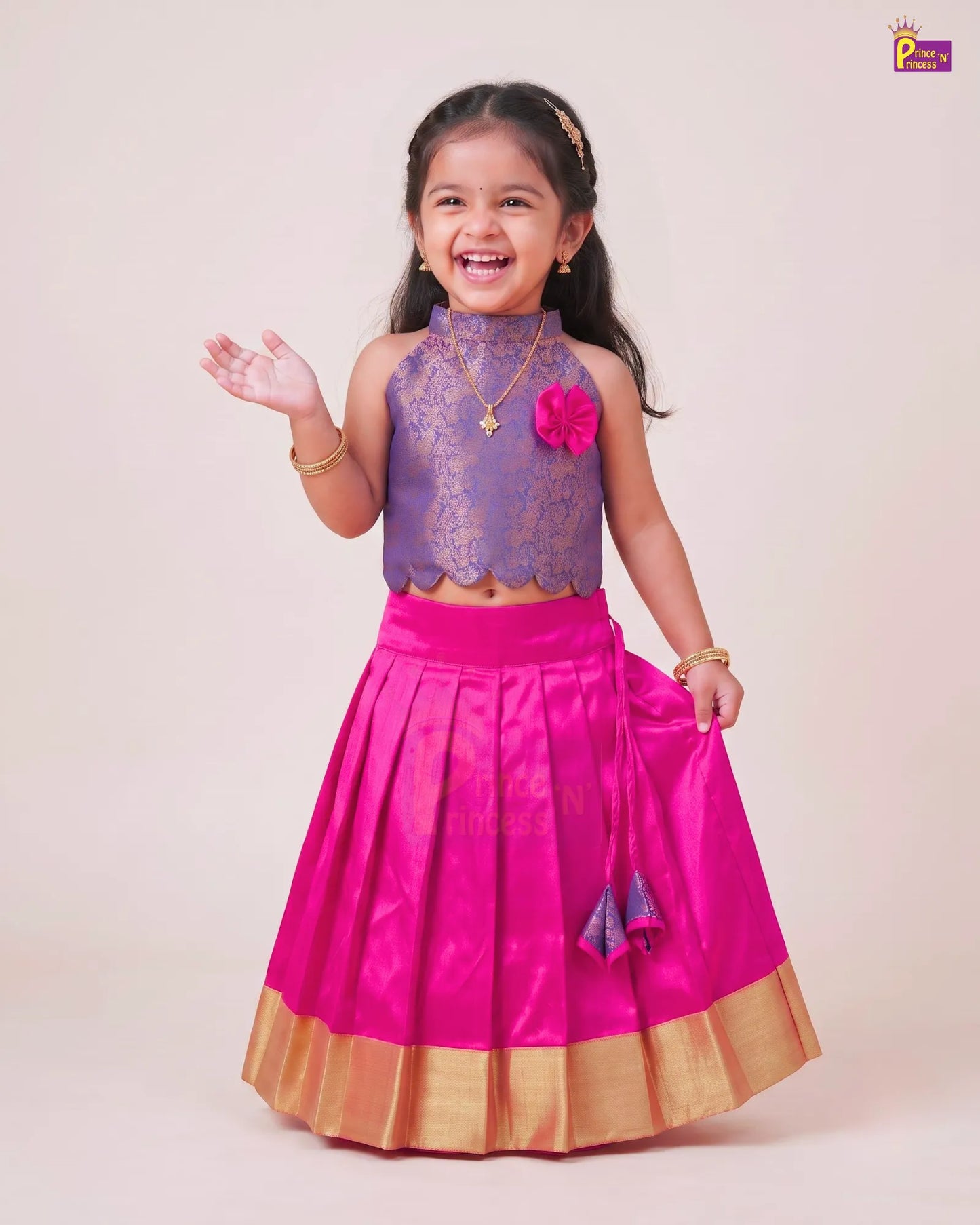 A young girl modeling a traditional Indian outfit consisting of a purple lace sleeveless top and a bright pink pleated skirt with a gold border, set against a plain beige studio background.