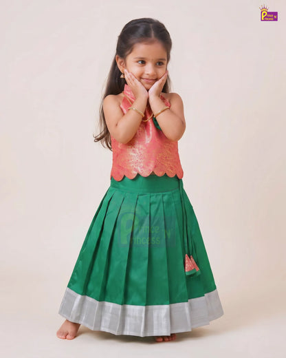 A young girl modeling a traditional Indian ethnic dress with a metallic pink sleeveless top and a pleated green skirt with a silver border, set against a plain beige background.