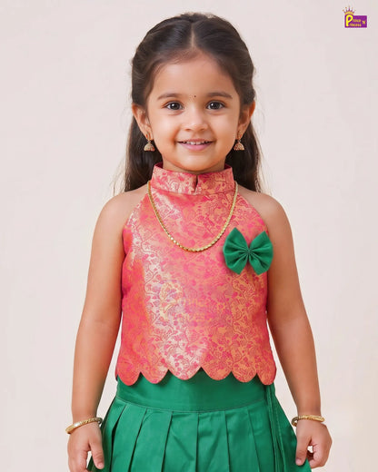 A young girl modeling a traditional Indian outfit consisting of a sleeveless coral pink and gold brocade top with a green bow accent, paired with a green skirt, set against a plain light beige background.