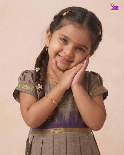 A young girl with braided hair smiles while posing with her hands on her cheeks, wearing a traditional Indian outfit in brown and purple with gold accents, set against a solid beige background.