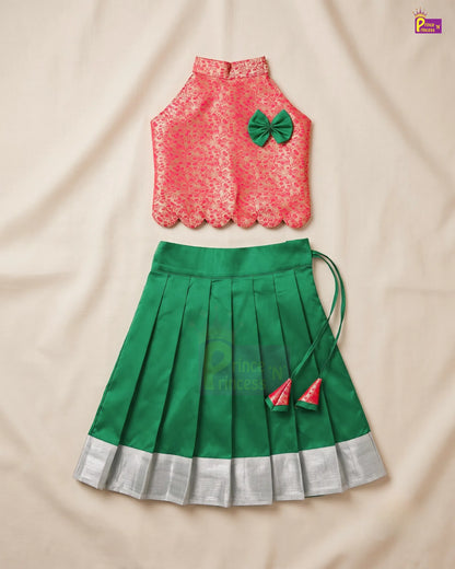 A two-piece girls' traditional outfit laid flat on a beige fabric background. The top is a sleeveless coral-pink lace blouse with scalloped edges and a green bow accent.