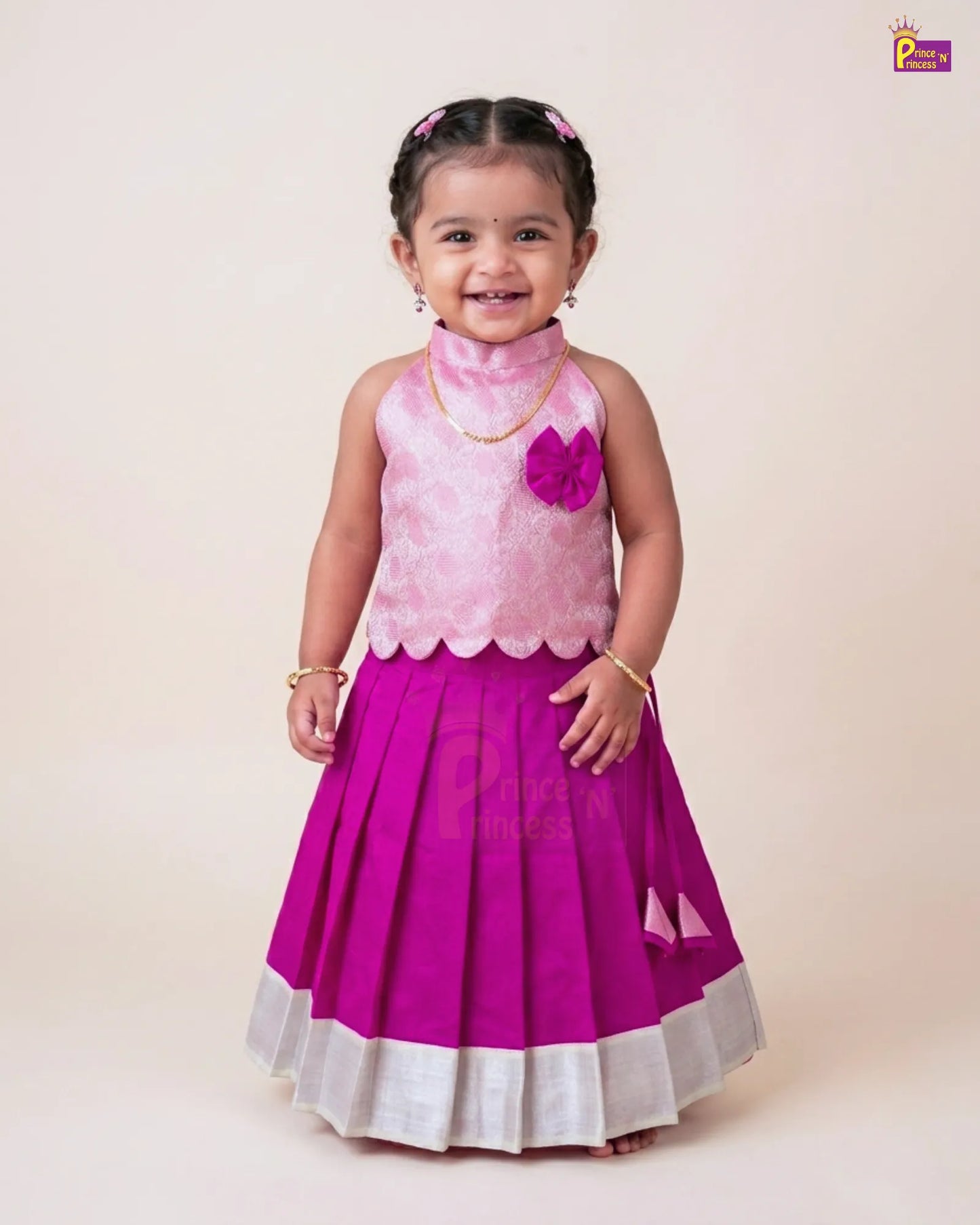 A young girl modeling a traditional Indian ethnic dress with a light pink textured sleeveless top and a bright magenta pleated skirt with a white border, set against a plain beige studio background.