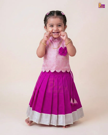 A young toddler girl wearing a traditional Indian outfit consisting of a light pink sleeveless top with scalloped edges and a bright magenta pleated skirt with a white border, standing against a plain beige background.