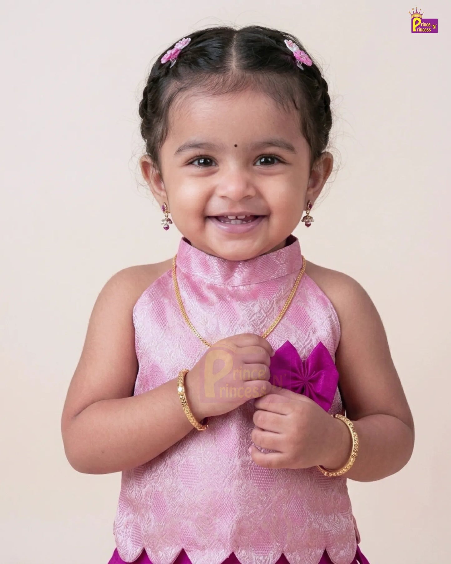 A young toddler girl smiling and wearing a pink traditional sleeveless top with a high collar, gold jewelry, and pink hair clips, photographed against a plain light beige background.