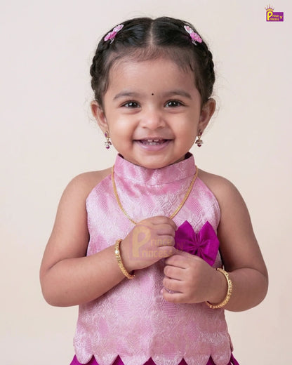 A young toddler girl smiling and wearing a pink traditional sleeveless top with a high collar, gold jewelry, and pink hair clips, photographed against a plain light beige background.