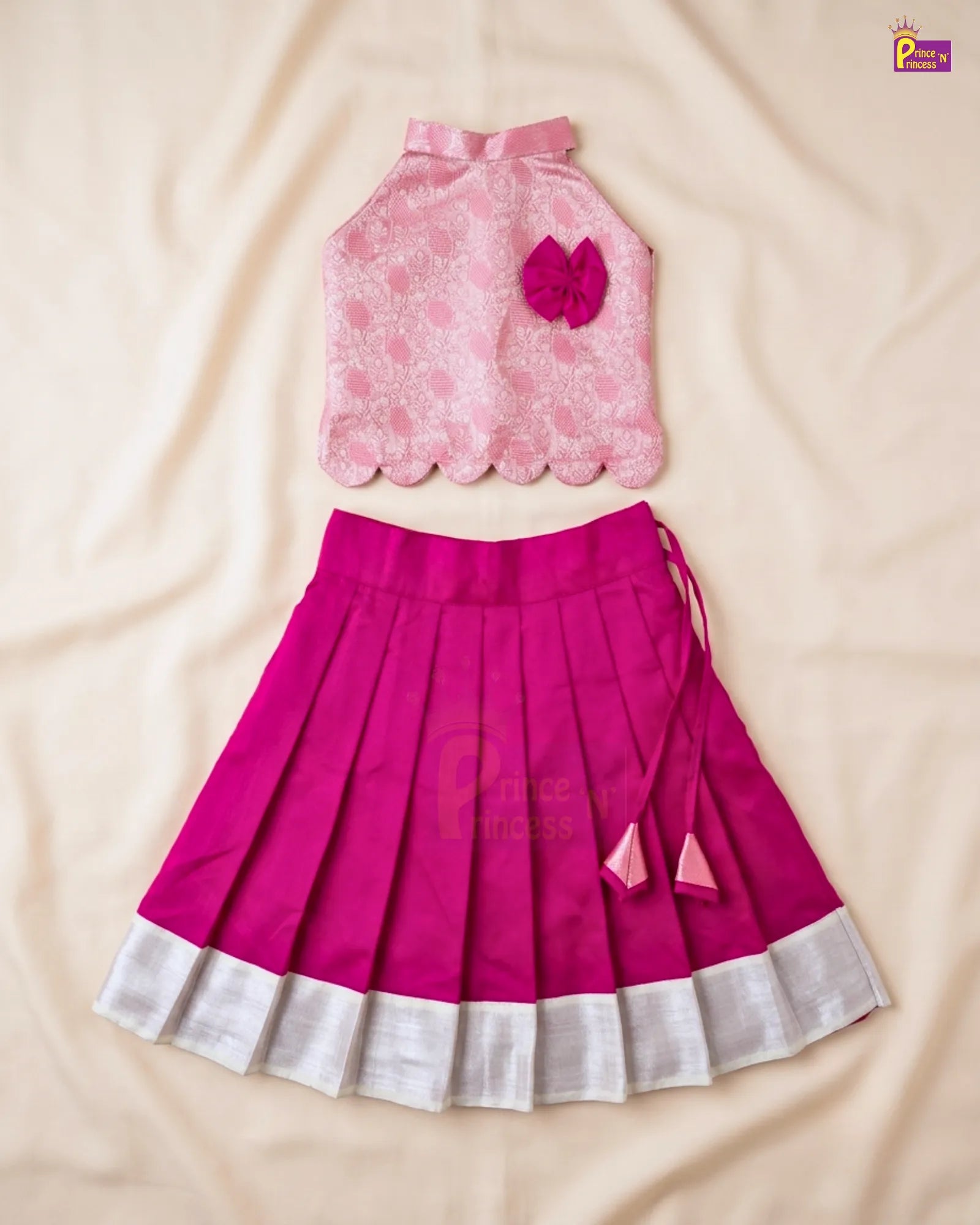 A two-piece girls' ethnic wear set featuring a pink floral lace sleeveless top with a scalloped hem and a bright magenta pleated skirt with a white ruffled hem, laid flat on a beige fabric background.