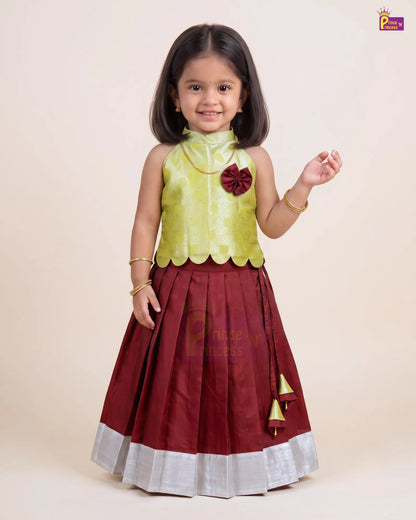 A young girl modeling a traditional Indian dress with a light green sleeveless top and a dark maroon pleated skirt with a silver border and gold tassel details, set against a plain beige background.