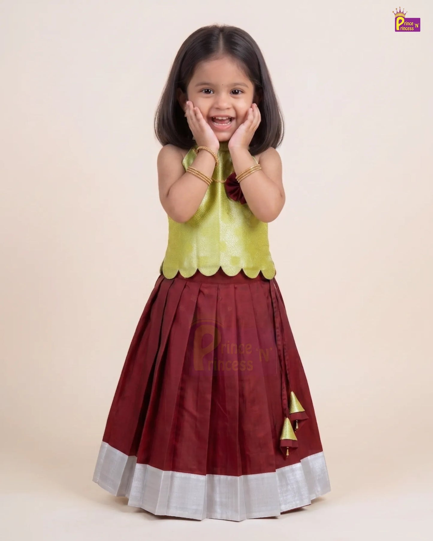 A young girl modeling a traditional Indian outfit consisting of a maroon skirt with a silver border and gold tassel detail, paired with a lime green sleeveless top, against a solid beige background.