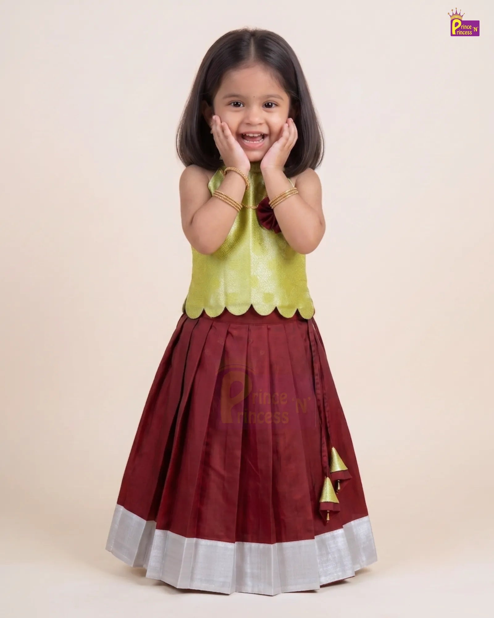 A young girl modeling a traditional Indian outfit consisting of a maroon skirt with a silver border and gold tassel detail, paired with a lime green sleeveless top, against a solid beige background.