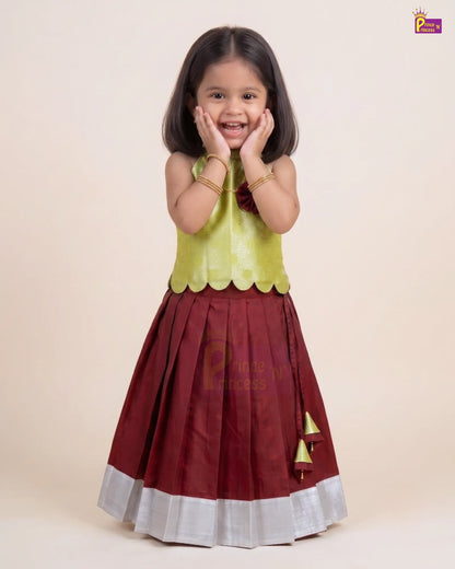A young girl modeling a traditional Indian outfit consisting of a maroon skirt with a silver border and gold tassel detail, paired with a lime green sleeveless top, against a solid beige background.