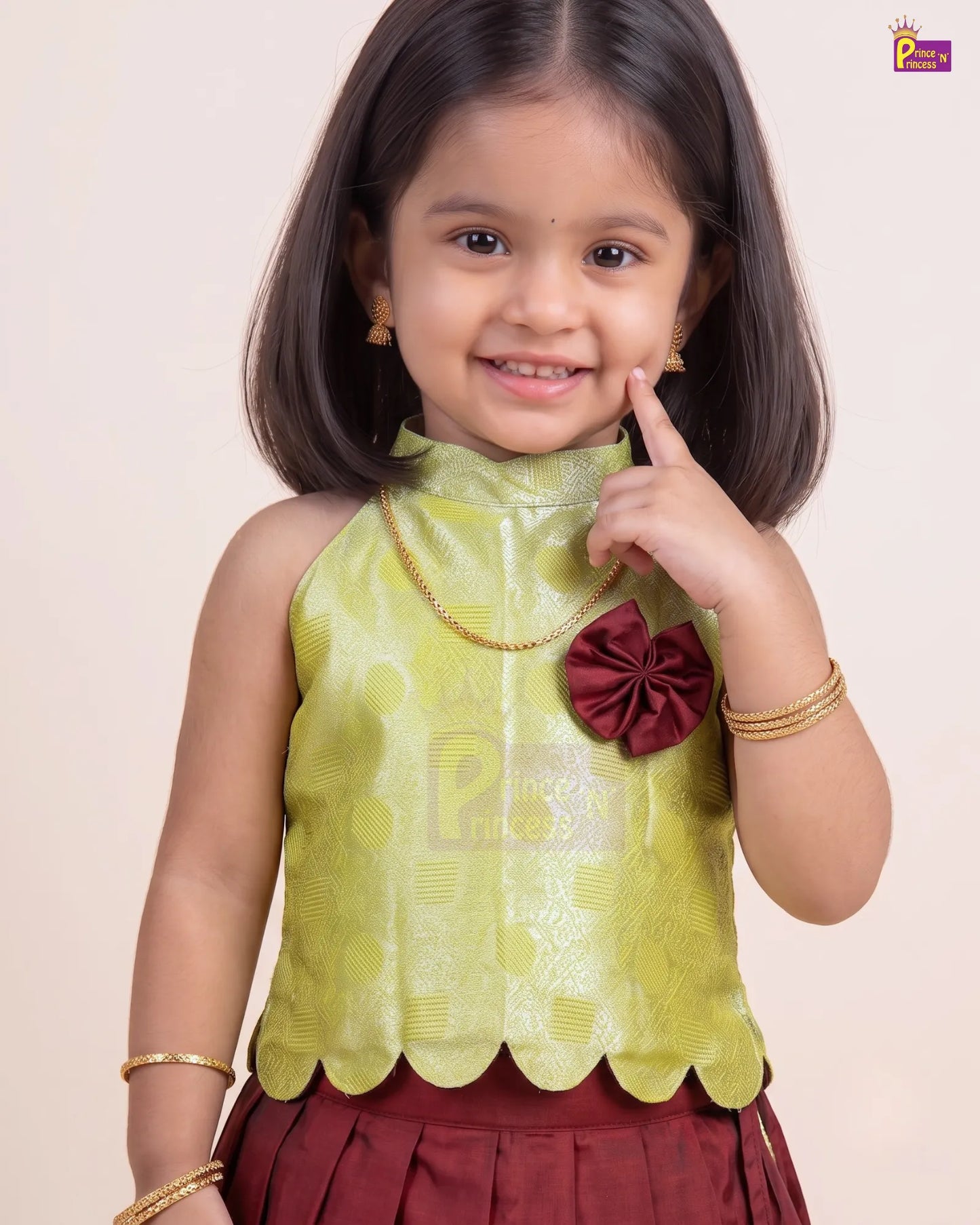 A young girl modeling a traditional Indian outfit featuring a lime green patterned sleeveless top with a scalloped hem and a maroon bow accent, paired with a maroon skirt and gold jewelry, set against a plain beige background.