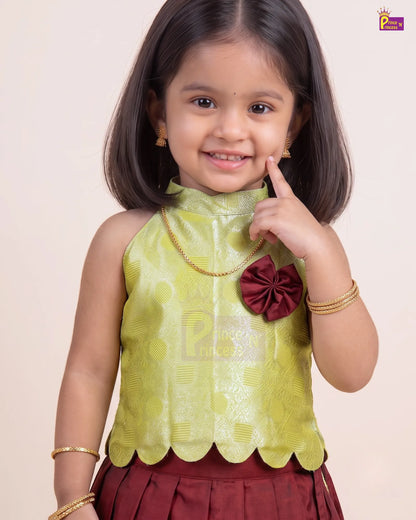 A young girl modeling a traditional Indian outfit featuring a lime green patterned sleeveless top with a scalloped hem and a maroon bow accent, paired with a maroon skirt and gold jewelry, set against a plain beige background.