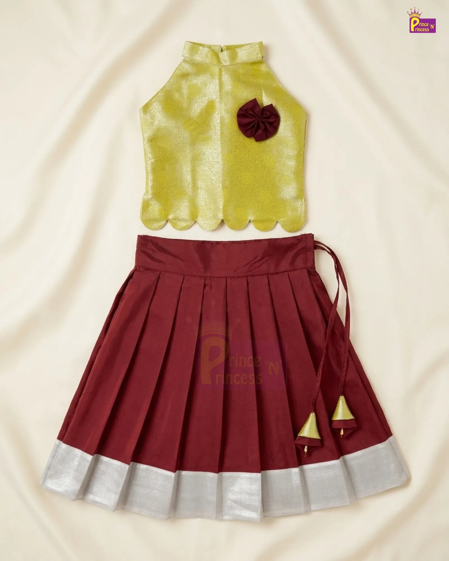 A two-piece girls' traditional outfit set consisting of a sleeveless lime green top with a dark red fabric flower accent and a pleated maroon skirt with a silver metallic hem and gold jhumka hanging ties, laid flat on a cream-colored fabric background.