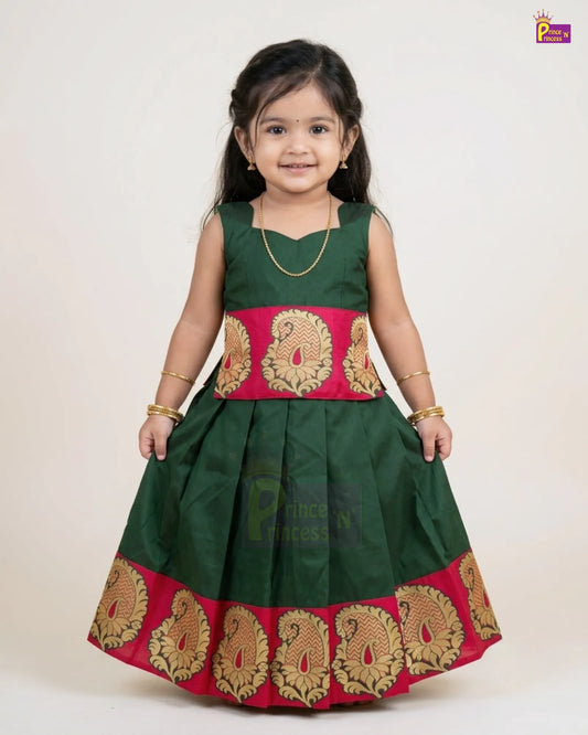 A young girl modeling a dark green traditional Indian dress with a wide red waistband and a matching red hem featuring ornate gold paisley borders. She is standing against a plain light beige background.