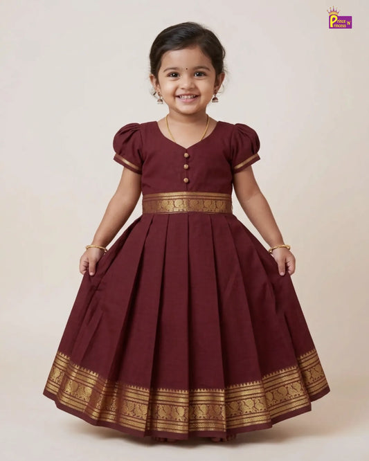 A young girl wearing a maroon A-line dress with gold zari borders and button details, standing against a plain beige background.