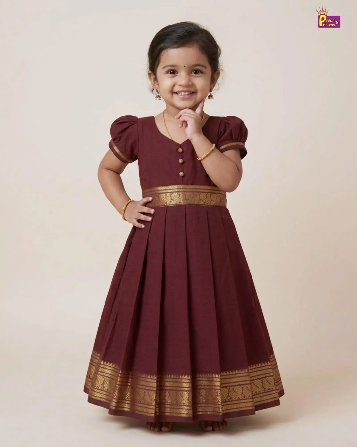 A young girl modeling a maroon A-line ethnic dress with gold zari borders and button details, set against a solid beige studio background.