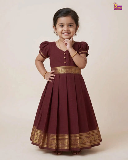 A young girl modeling a maroon A-line ethnic dress with gold zari borders and button details, set against a solid beige studio background.