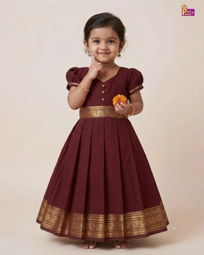 A young girl modeling a traditional maroon A-line dress with gold zari borders and button details, standing against a solid beige studio background.