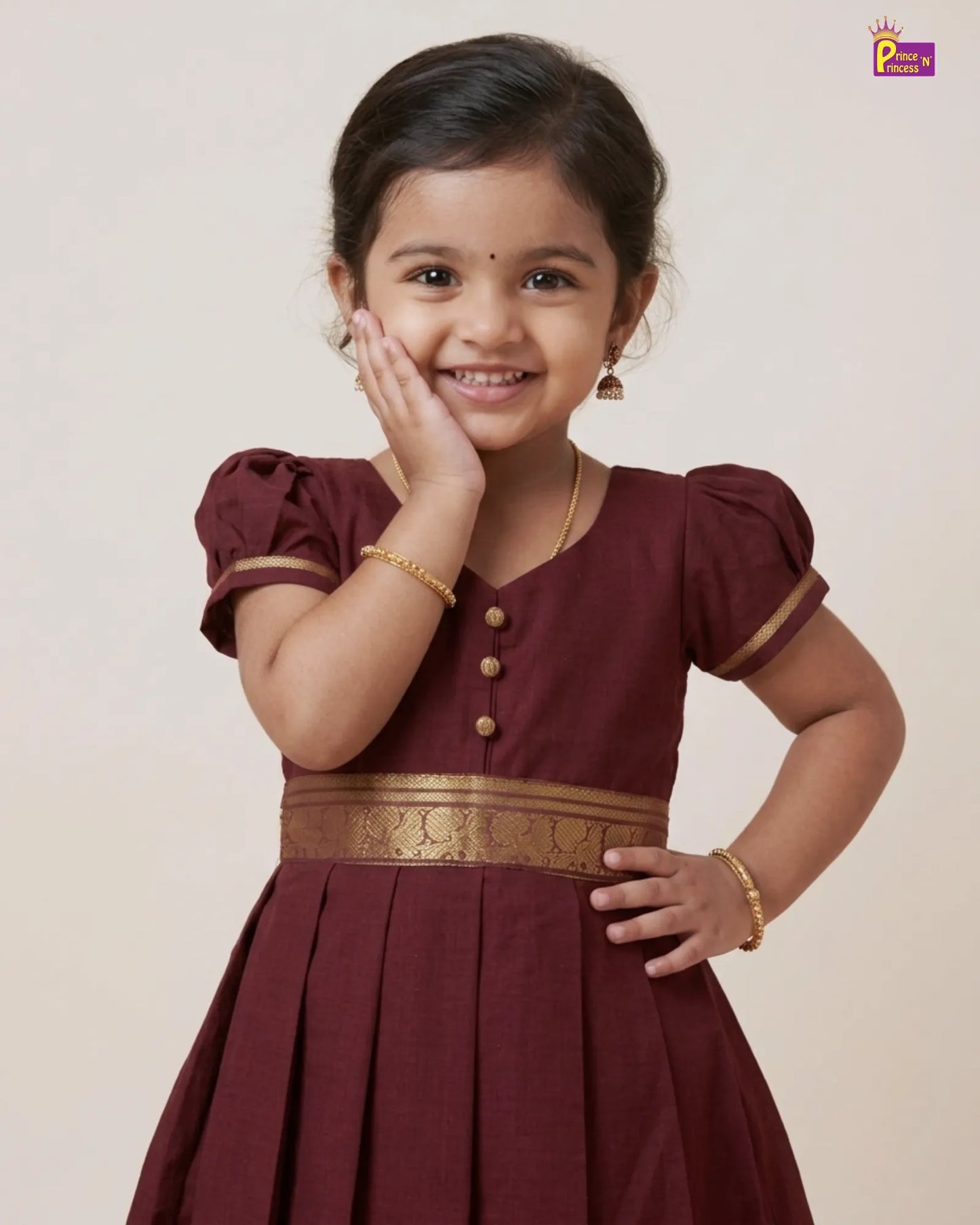 A young girl smiling and posing in a maroon traditional dress with gold trim and a gold patterned waistband, set against a plain beige background.