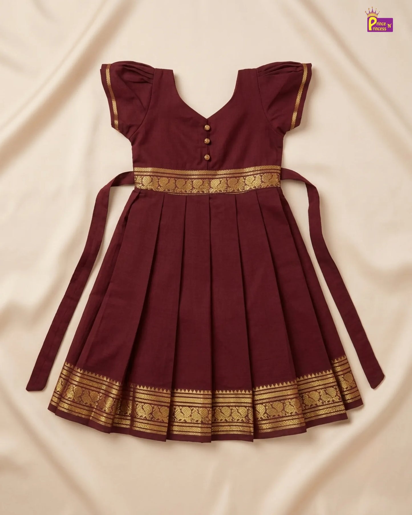 A maroon pleated baby dress with gold patterned borders and a matching tie belt, laid flat on a cream-colored fabric background.