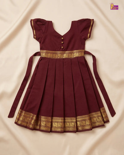 A maroon pleated baby dress with gold patterned borders and a matching tie belt, laid flat on a cream-colored fabric background.