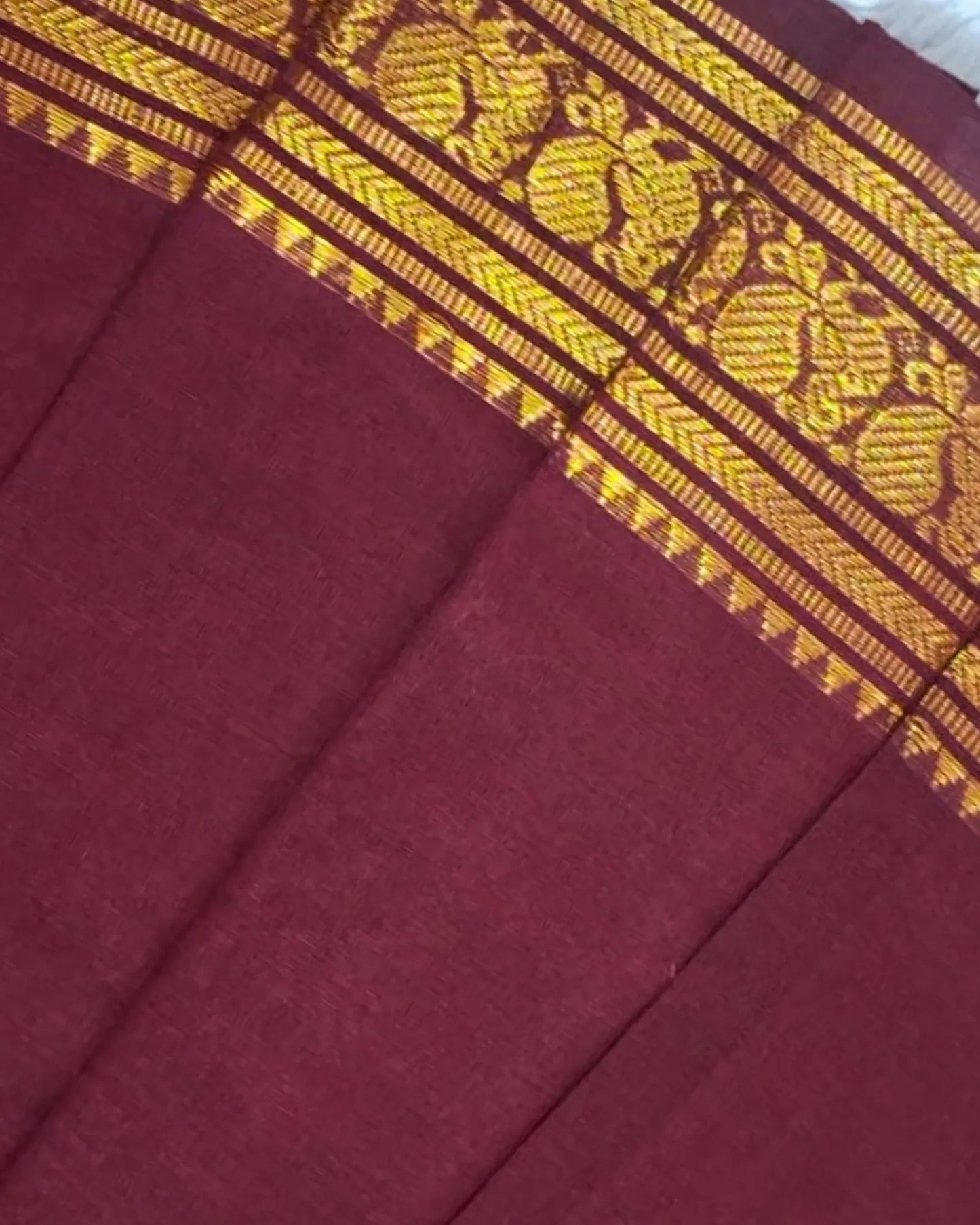 A close-up of a deep maroon fabric with a wide ornate gold woven border featuring traditional motifs.