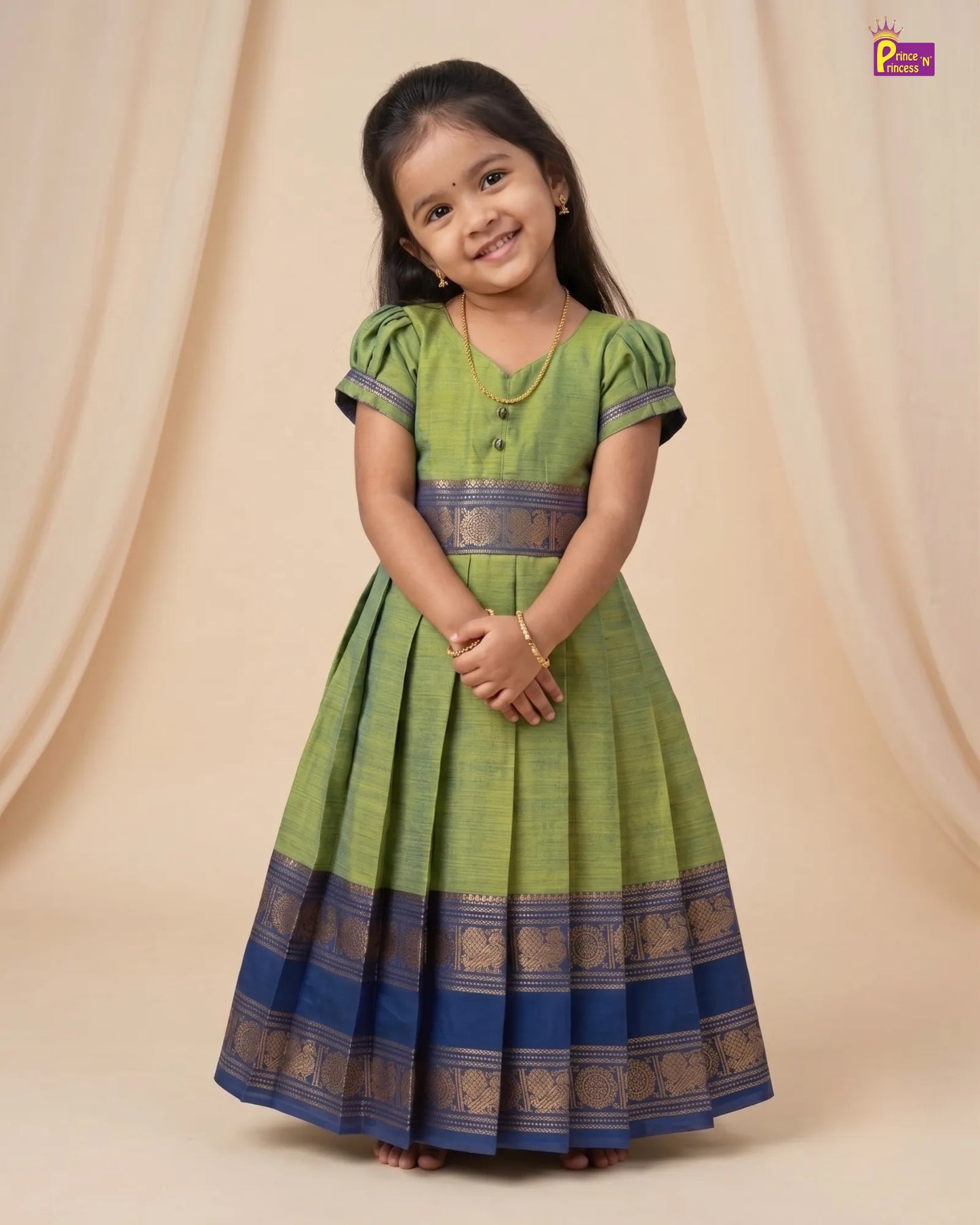 A young girl modeling a green and blue traditional Indian dress with gold zari borders, set against a beige draped fabric backdrop.