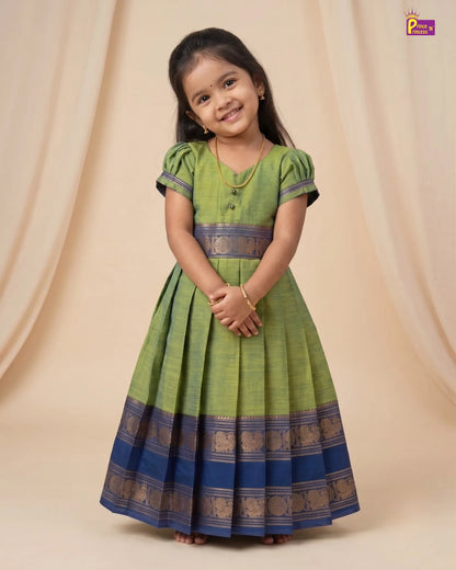 A young girl modeling a green and blue traditional Indian dress with gold zari borders, set against a beige draped fabric backdrop.