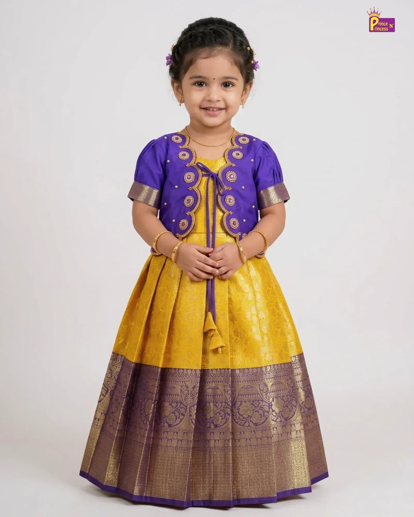 A young girl modeling a traditional Indian ethnic outfit consisting of a yellow and purple kurta with gold zari embroidery, set against a plain light gray background.