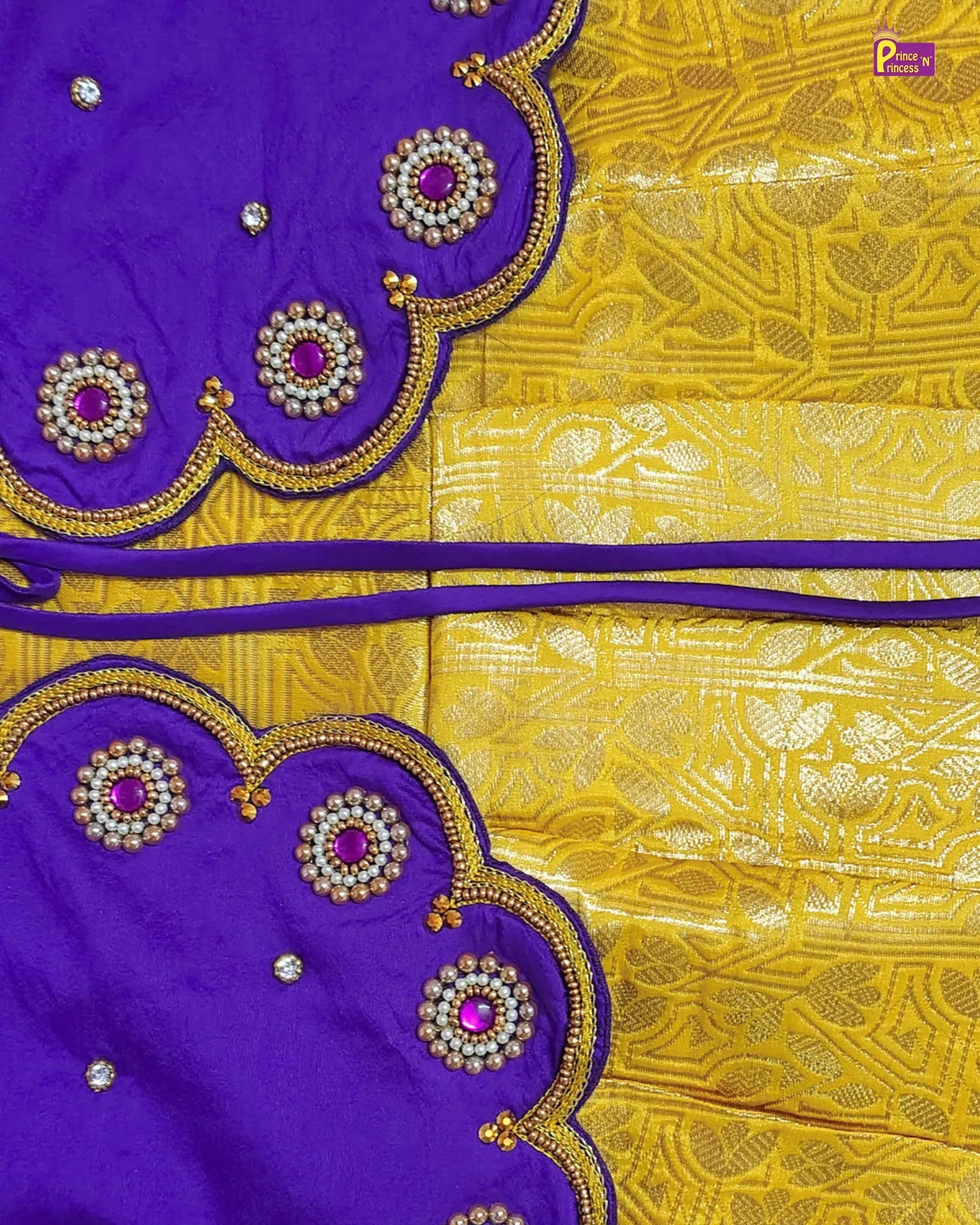 A close-up of a purple and yellow fabric textile, likely a saree or scarf, featuring a richly patterned yellow body with metallic gold motifs and matching purple borders decorated with scalloped edges, gold embroidery, and pink gemstone accents.