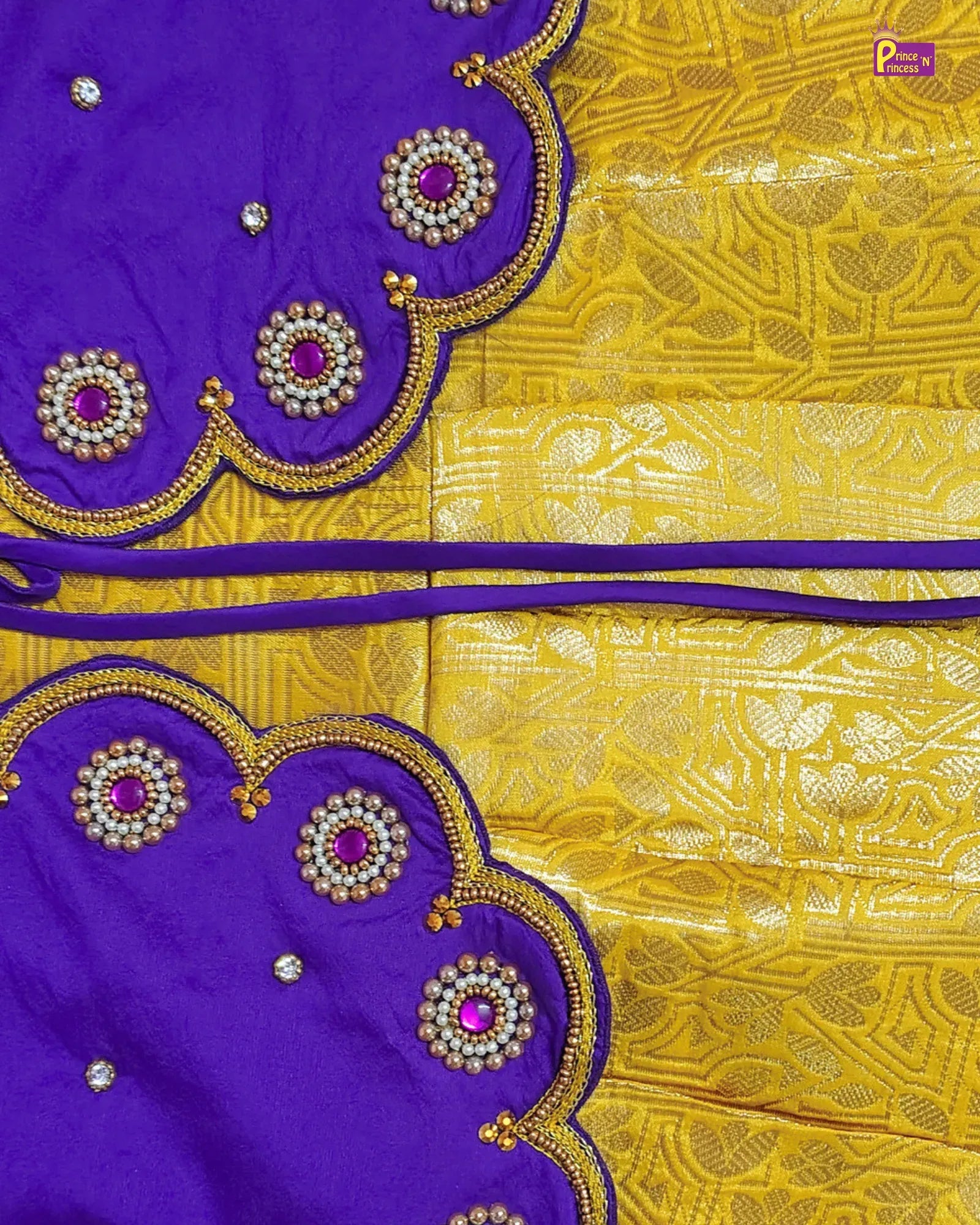 A close-up of a purple and yellow fabric textile, likely a saree or scarf, featuring a richly patterned yellow body with metallic gold motifs and matching purple borders decorated with scalloped edges, gold embroidery, and pink gemstone accents.
