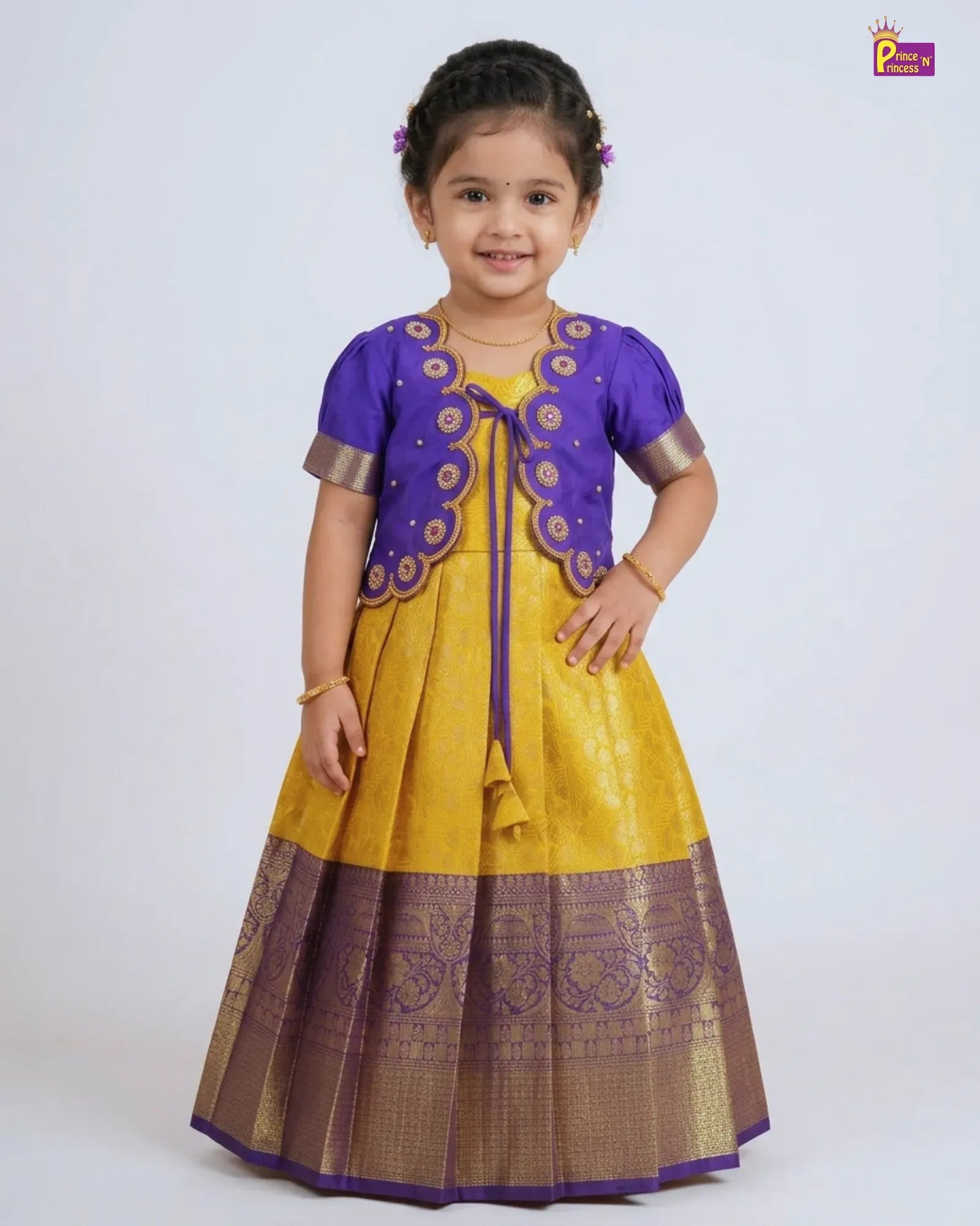 A young girl modeling a traditional Indian ethnic dress consisting of a yellow skirt with a wide purple and gold border, a matching purple embroidered vest, and a yellow blouse with purple sleeves.