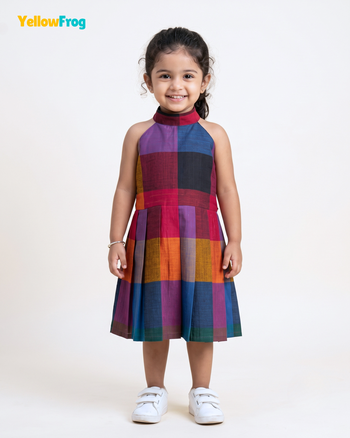 A young girl stands against a plain white studio background wearing a sleeveless high-neck dress with a colorful patchwork plaid pattern in purple, red, orange, blue, green, and black. She is wearing white sneakers, and the YellowFrog logo is visible in the top left corner.