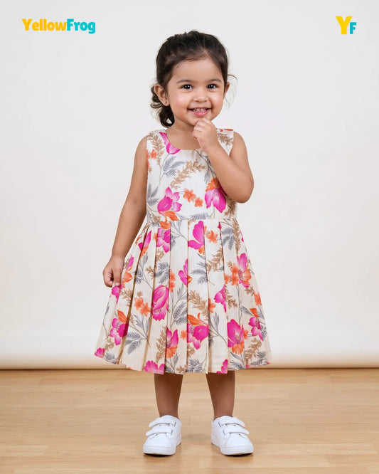 A young girl stands in a studio wearing a sleeveless pleated dress with a colorful floral print on a cream background, paired with white shoes. The YellowFrog brand logo is visible in the top left corner.