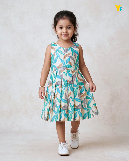 A young girl wearing a sleeveless pleated dress with a colorful leaf pattern in teal, brown, and beige, paired with white sneakers, standing against a textured beige backdrop.