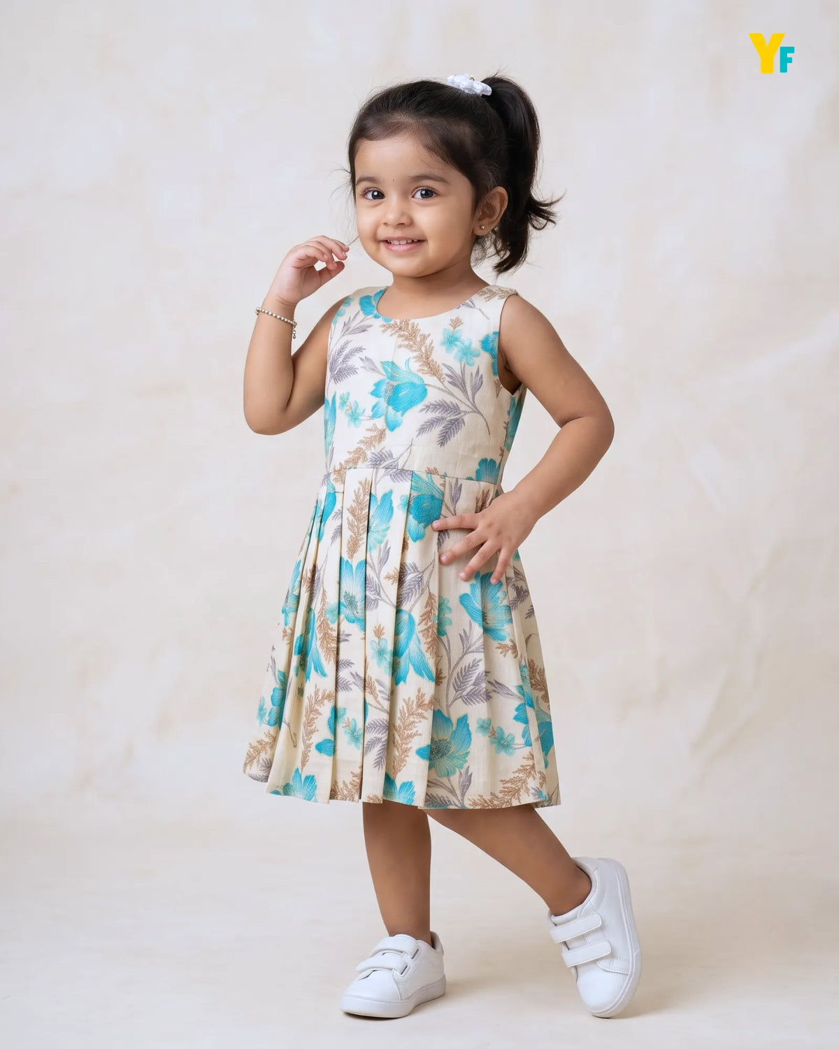 A young girl with dark hair in a ponytail wearing a sleeveless pleated dress with a blue and beige floral pattern, paired with white sneakers, posing against a light beige textured backdrop.