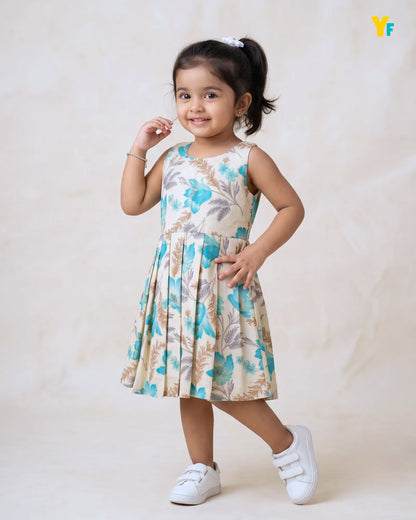A young girl with dark hair in a ponytail wearing a sleeveless pleated dress with a blue and beige floral pattern, paired with white sneakers, posing against a light beige textured backdrop.
