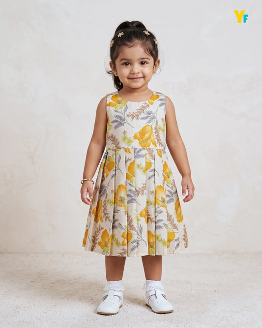A young girl with her hair in a ponytail stands wearing a sleeveless pleated dress with a yellow and green floral pattern on a cream background. She is wearing white shoes and standing against a textured light beige wall.