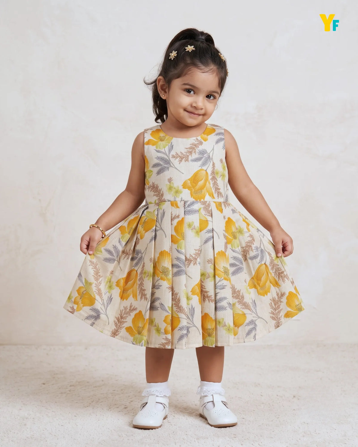 A young girl with her hair in a high ponytail wearing a sleeveless, pleated dress with a yellow and gray floral print on a cream background. She is standing against a textured beige wall and floor, wearing white shoes and socks.