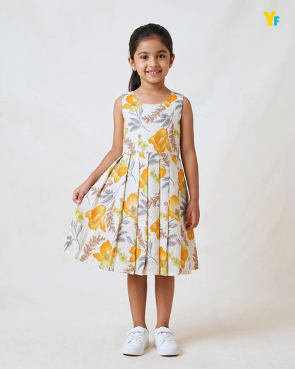 A young girl stands against a plain light background wearing a sleeveless pleated dress with a yellow and gray botanical print, paired with white sneakers.