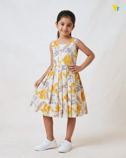 A young girl modeling a sleeveless pleated dress with a yellow and gray floral print on a cream background, paired with white sneakers, against a plain light beige studio backdrop.