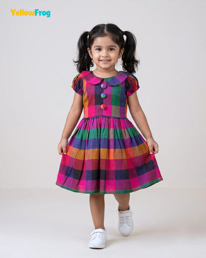 A young girl with dark hair in pigtails models a colorful plaid dress with pink, green, orange, and purple stripes, featuring a buttoned collar and puff sleeves. She is standing against a plain light gray background, with the YellowFrog logo in the top left corner.