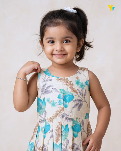 A young girl with dark hair in a ponytail wearing a sleeveless cream-colored dress with a blue and brown floral pattern, posing against a light beige background.