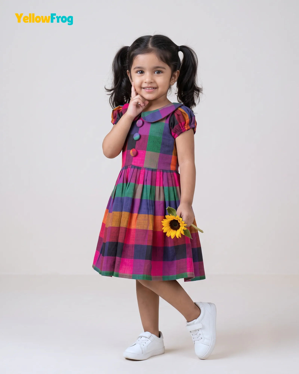 A young girl with dark hair in pigtails models a colorful plaid dress with puff sleeves and a buttoned yoke, holding a small yellow sunflower. She is standing against a plain light gray studio background.
